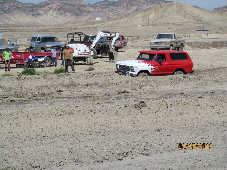 Red and white Ford Bronco stuck in mud, with other vehicles and people around