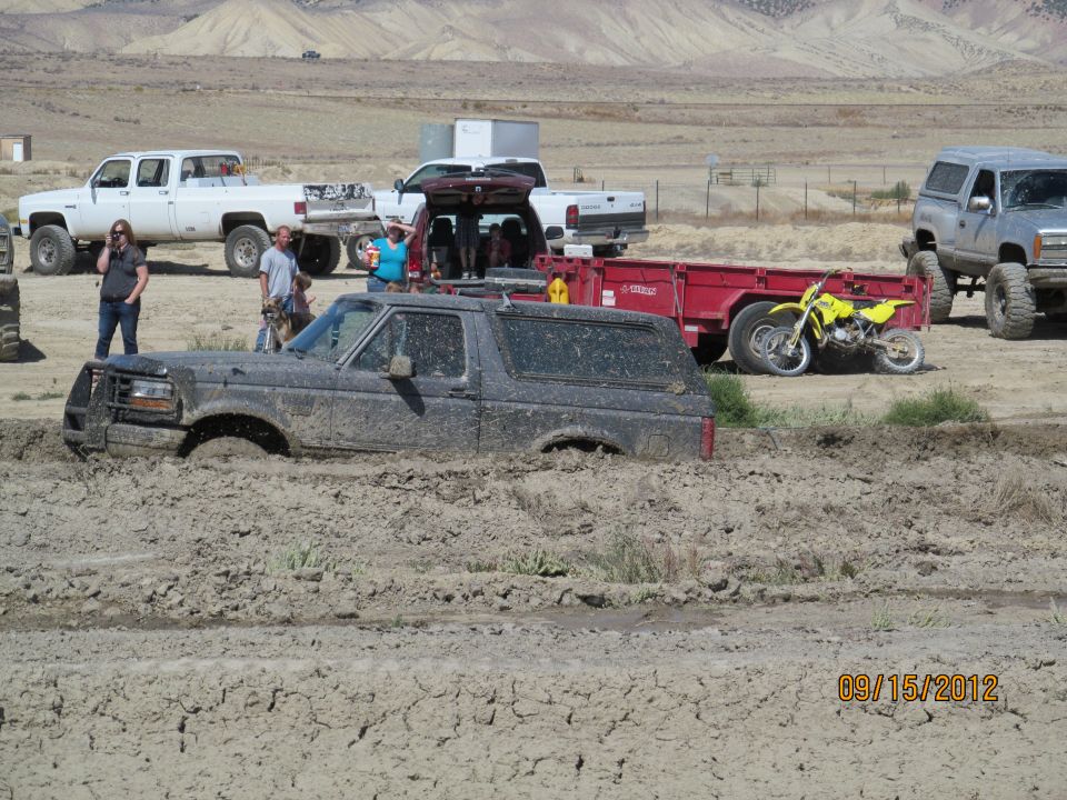 Mud-covered 1995 Ford Bronco stuck in deep mud, with other trucks and a dirt bike visible in the background