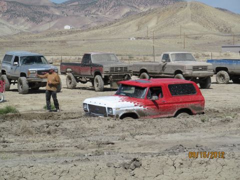 1979 Ford Bronco - Multiple off-road vehicles are seen in a muddy terrain, with one red and white Ford Bronco partially submerged.