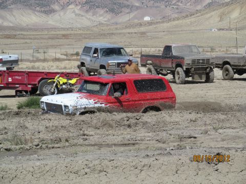 1979 Ford Bronco - A red and white Ford Bronco is stuck deep in the mud during a mud bog event, with other vehicles and a dirt bike visible in the background.