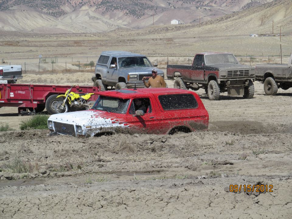 Red and white 1978 Ford Bronco stuck in mud, with other trucks and a motorcycle nearby