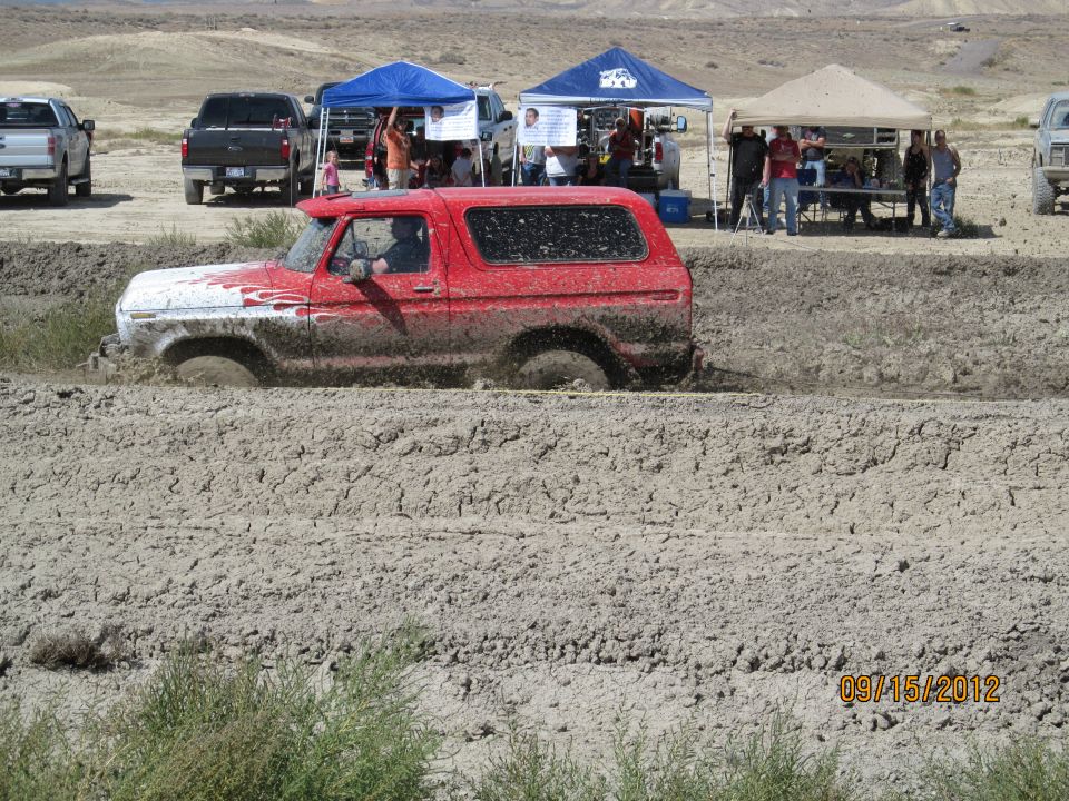 Red 1979 Ford Bronco driving through a mud pit with other vehicles and tents in the background