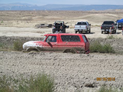 1979 Ford Bronco - A red SUV is stuck deep in mud, with other vehicles and a trailer in the background.