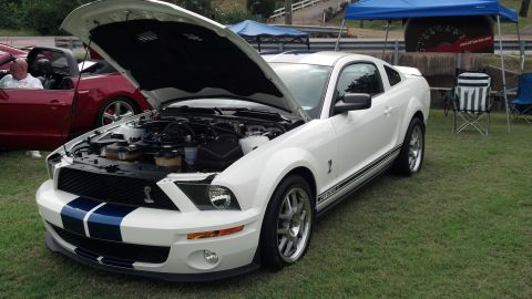 2007 Ford Mustang - A white Ford Mustang Shelby GT500 is parked on grass with its hood open, showing the engine. Another car is visible in the background.