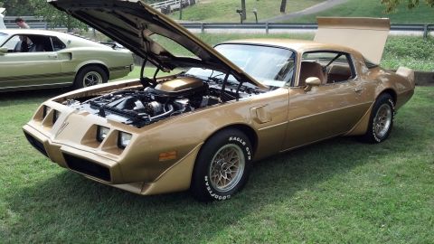 2007 Ford Mustang - A gold 1977 Pontiac Firebird is displayed with its hood open, revealing the engine. Another car is visible in the background.