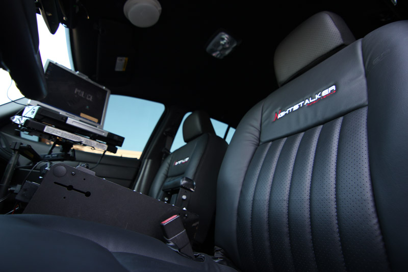 2004 Ford Crown Victoria police car interior showing front seats, console with laptop, and 'GHOSTALKER' embroidery on passenger seat