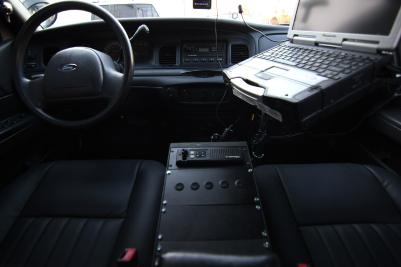Interior of a 2004 Ford Crown Victoria police car, showing the steering wheel, dashboard, laptop, and radio equipment
