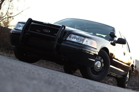 2004 Ford Crown Victoria - A black Ford Crown Victoria with a push bar is angled on a road with a bright sky.