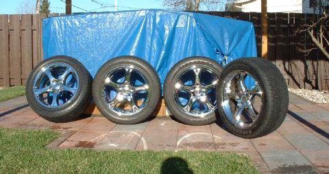 2000 Ford Crown Victoria - Four chrome wheels and tires are displayed on a paved patio area in front of a blue tarp and wooden fence.