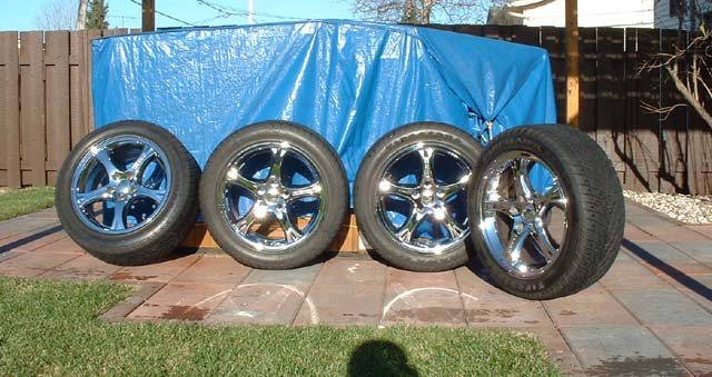 Four chrome five-spoke wheels with tires lined up on a patio in front of a blue tarp