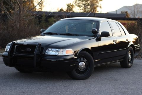 2004 Ford Crown Victoria - A black 2004 Ford Crown Victoria police car is parked on a paved surface with trees and a building in the background.