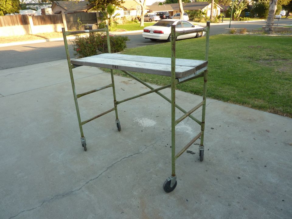 Rolling scaffolding unit with wooden planks on a concrete driveway. A white car is parked in the background