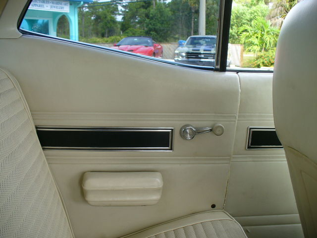 Interior of a 1970 Ford Torino showing a cream door panel, chrome handle, and armrest, with a red car and a blue Ford Torino visible through the window