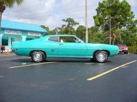 1970 Ford Torino - Side profile of a turquoise 1970 Ford Torino coupe parked in a lot with a building and trees.