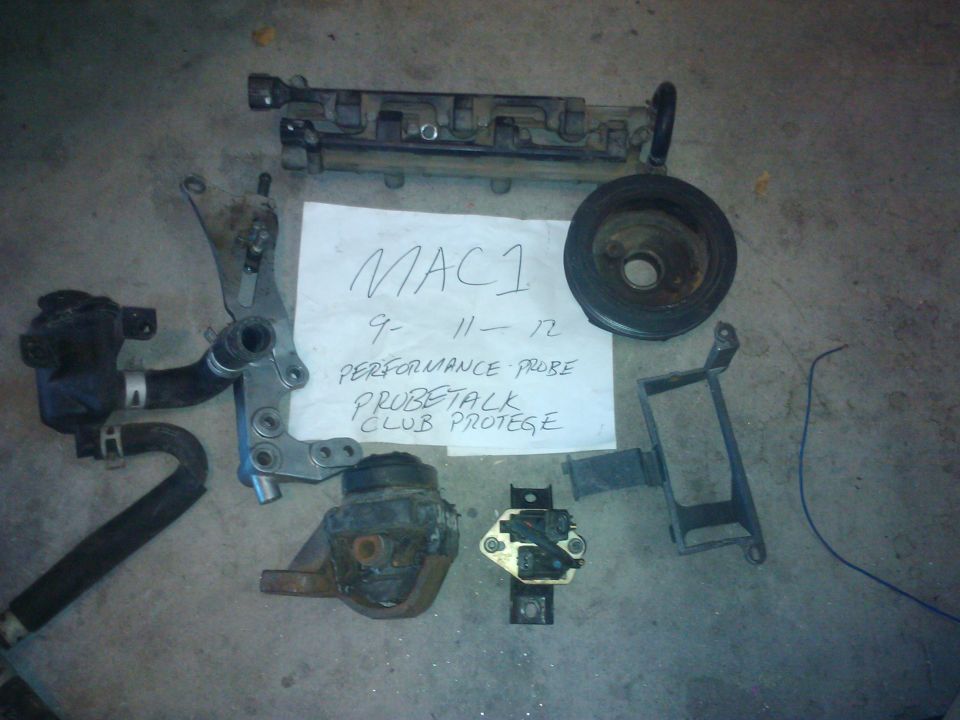 Engine parts including a pulley, manifold, and mounts laid out on concrete