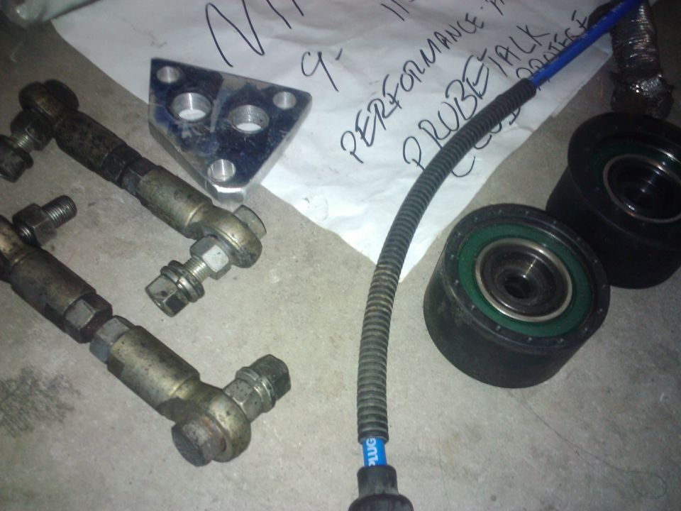 Several car parts including tie rods, a metal bracket, and pulleys laid out on the floor. Paper with writing is in the background