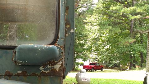 1978 Ford Bronco - A close-up view of a weathered green truck door and side mirror, with a red pickup truck visible in the distance.