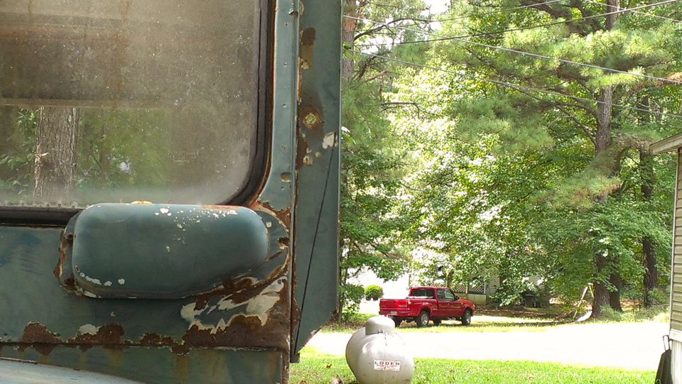 Close-up of rusty green truck door and side mirror; red pickup truck in background