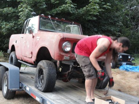 1961 International Scout 80 - A person works on a red off-road vehicle being transported on a trailer, with another vehicle in the background.