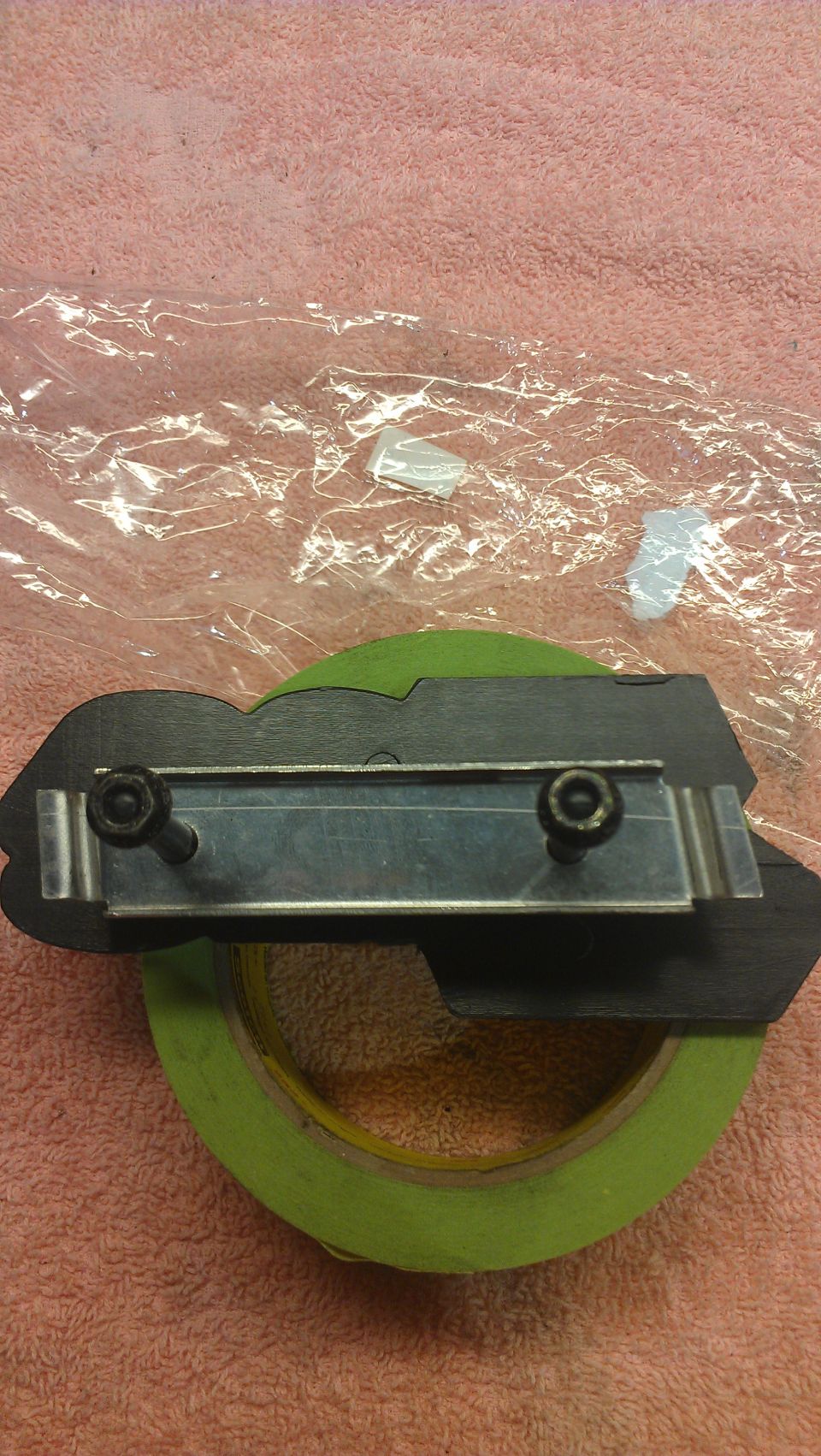 Black automotive emblem with two screws attached to a metal bracket, resting on green tape