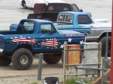 1989 Ford Bronco - A lifted blue pickup truck with patriotic graphics is parked next to another truck in an outdoor lot.