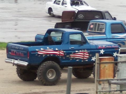 1989 Ford Bronco - A lifted blue Ford Bronco with patriotic decals is parked behind other vehicles.