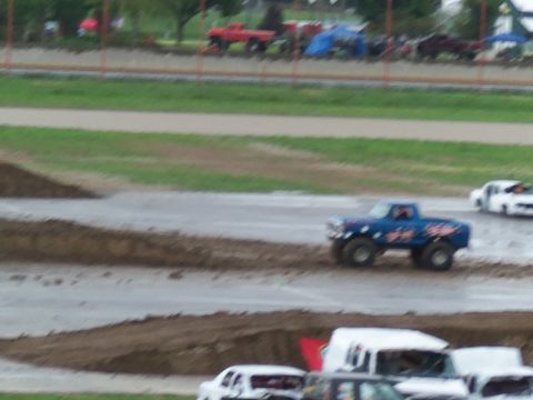 1989 Ford Bronco - A blue monster truck with custom graphics drives across a dirt track, with other vehicles visible in the background.