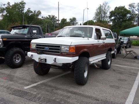 1989 Ford Bronco - A lifted Ford Bronco and a Ford pickup truck are parked in a lot under a cloudy sky.