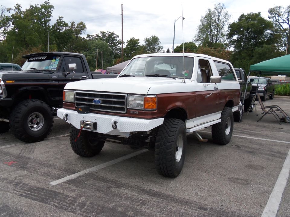 White and brown Ford Bronco with lifted suspension and aftermarket bumper. Black Ford pickup truck parked to the left