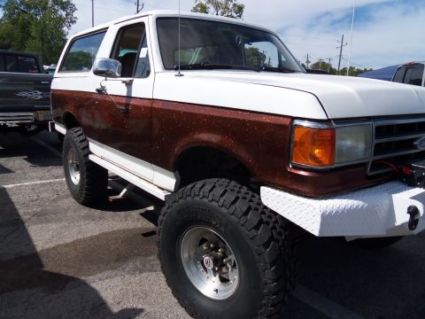 1989 Ford Bronco - A lifted 1989 Ford Bronco with a two-tone paint job sits in a parking lot under a partly cloudy sky.
