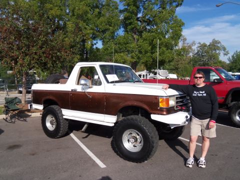 1989 Ford Bronco - A lifted 1989 Ford Bronco in white and brown is parked next to a man, with a red truck in the background.