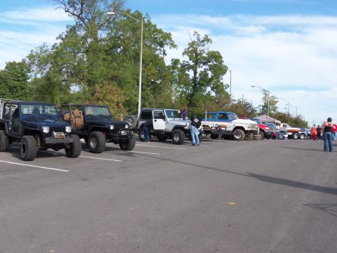 1989 Ford Bronco - A line of customized off-road vehicles are parked in a lot on a sunny day.