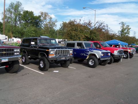 1989 Ford Bronco - A row of lifted trucks and SUVs are parked in a lot under a blue sky, with a black Ford Bronco in the foreground.