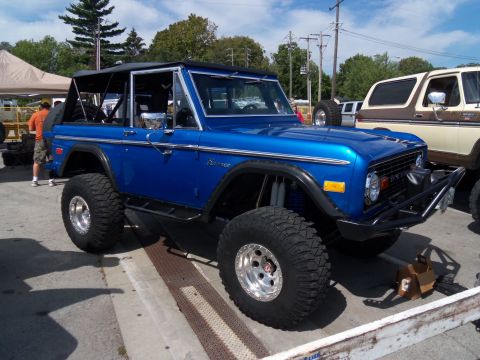 1989 Ford Bronco - This lifted blue Ford Bronco features a black soft top and aggressive off-road tires, parked next to another vintage SUV.