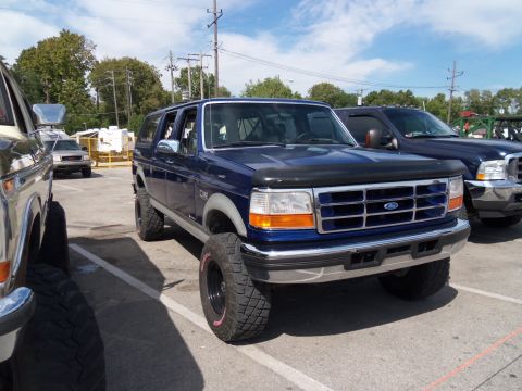 1989 Ford Bronco - A lifted blue Ford Bronco is parked in a lot next to other vehicles under a partly cloudy sky.