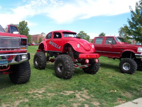 1989 Ford Bronco - A red monster truck with "BUGSZILLA" written on its side is parked on a grassy field next to other vehicles.