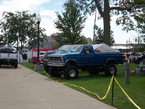 1989 Ford Bronco - A lifted blue pickup truck is parked on the grass at an outdoor event.