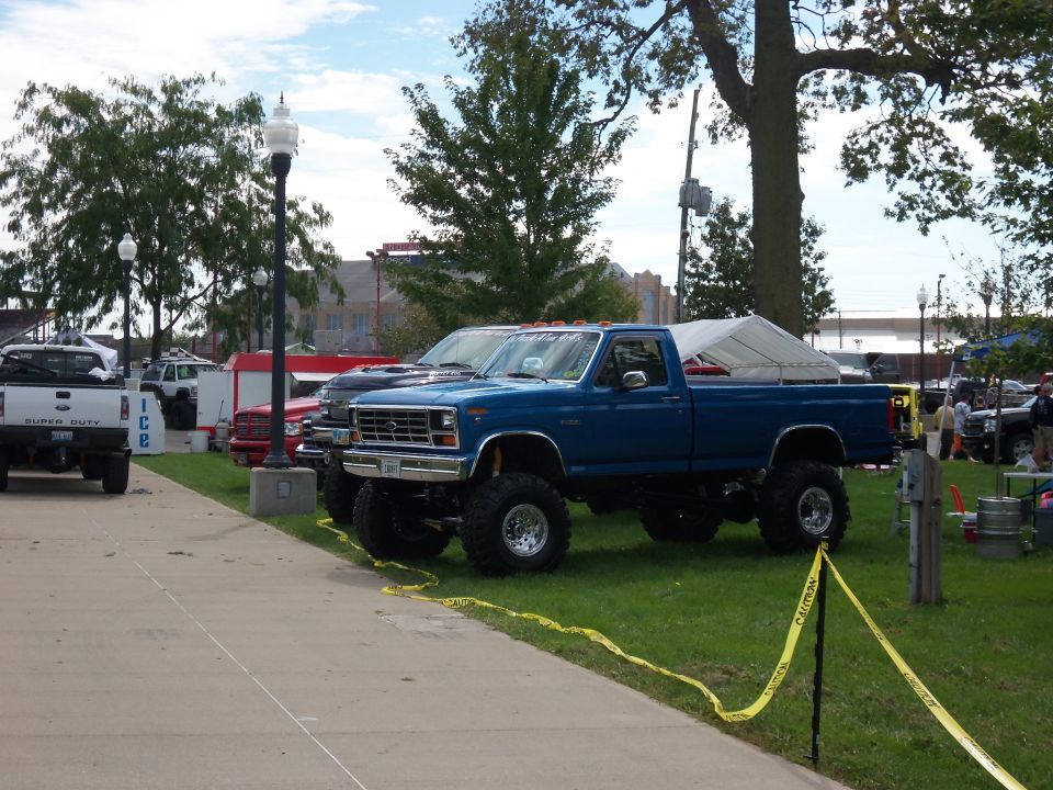 Blue lifted pickup truck with a white tarp over the bed, parked on grass next to a sidewalk. Other vehicles are visible in the background