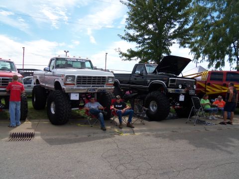 1989 Ford Bronco - Two custom lifted pickup trucks are displayed outdoors with people relaxing around them.
