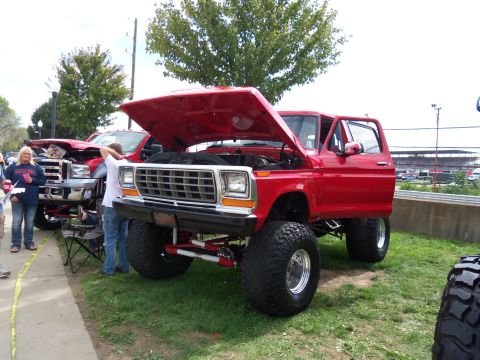 1989 Ford Bronco - A lifted red Ford Bronco from the late 1970s is displayed with its hood and door open, parked next to another truck.