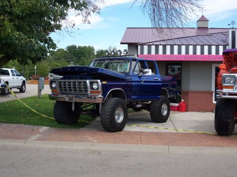 1989 Ford Bronco - This lifted blue Ford truck, likely from the 1970s, is parked next to a white truck outside a building with pink signage.