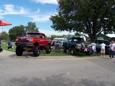 1989 Ford Bronco - Two heavily modified pickup trucks are displayed outdoors under a large tree.