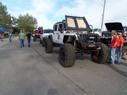 1989 Ford Bronco - Several modified off-road vehicles are lined up on a street, with people milling around.