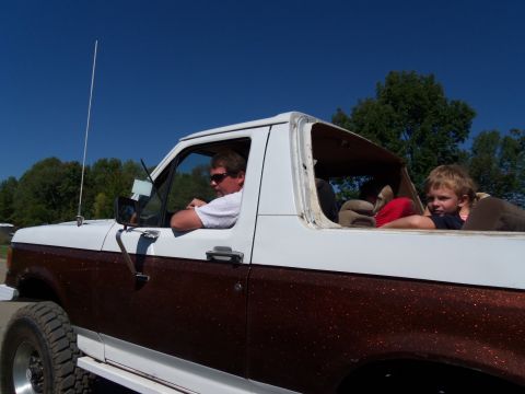 1989 Ford Bronco - A man drives a two-tone Ford Bronco with a child riding in the rear passenger area.