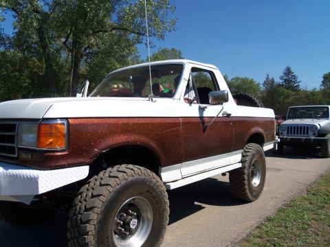 1989 Ford Bronco - A lifted 1989 Ford Bronco with a two-tone paint job is seen on a road, with another Jeep following behind.