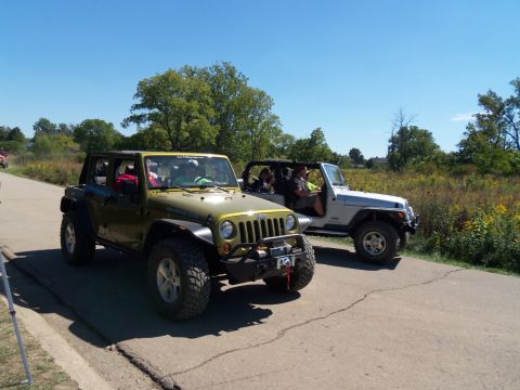 1989 Ford Bronco - Two Jeep Wranglers drive down a road on a sunny day, with trees and tall grass on the side.