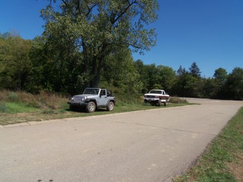 1989 Ford Bronco - A silver Jeep Wrangler and a white pickup truck are parked on the side of a road on a clear day.