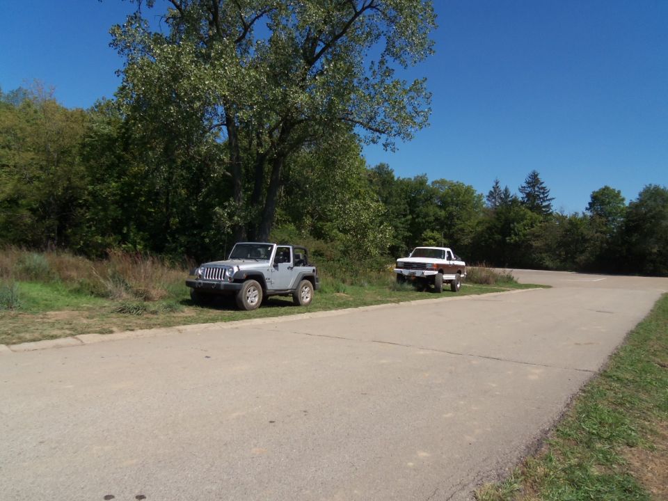 Silver Jeep Wrangler and white pickup truck parked on a grassy shoulder under a large tree