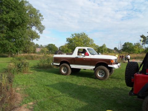 1989 Ford Bronco - A lifted Ford Bronco with a two-tone paint job sits in a field, with a group of people in the background.
