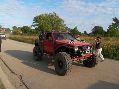 1989 Ford Bronco - A modified red SUV with a roll cage and oversized tires is parked on a road next to a field.